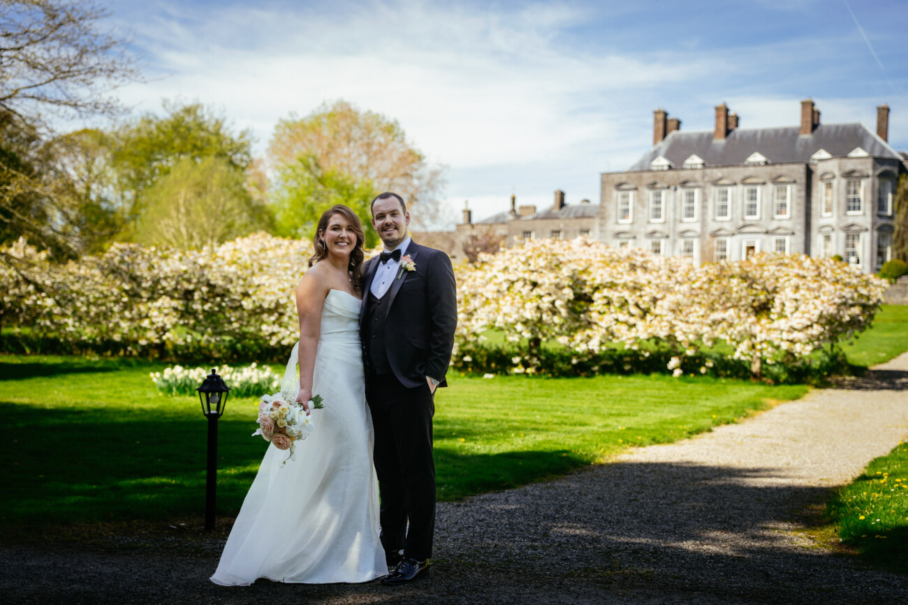 Bride and groom pose together outdoors on a sunny day, standing on a gravel path with a historic building and blooming shrubs in the background—a perfect moment captured through the elegance of Castle Durrow Wedding Photography.