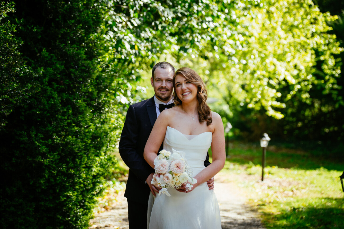 A bride in a strapless white gown holds a bouquet and stands in front of a groom in a dark suit, both smiling outdoors in a sunlit, green garden—perfectly capturing the elegance and romance of Castle Durrow Wedding Photography.
