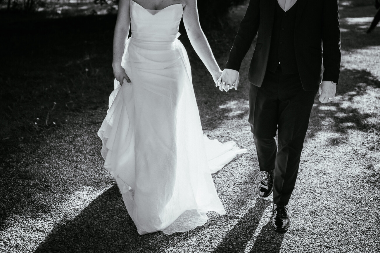 A couple in formal attire, one in a strapless white dress and the other in a suit, walk hand in hand outdoors on a gravel path—an elegant moment beautifully captured by Castle Durrow Wedding Photography.