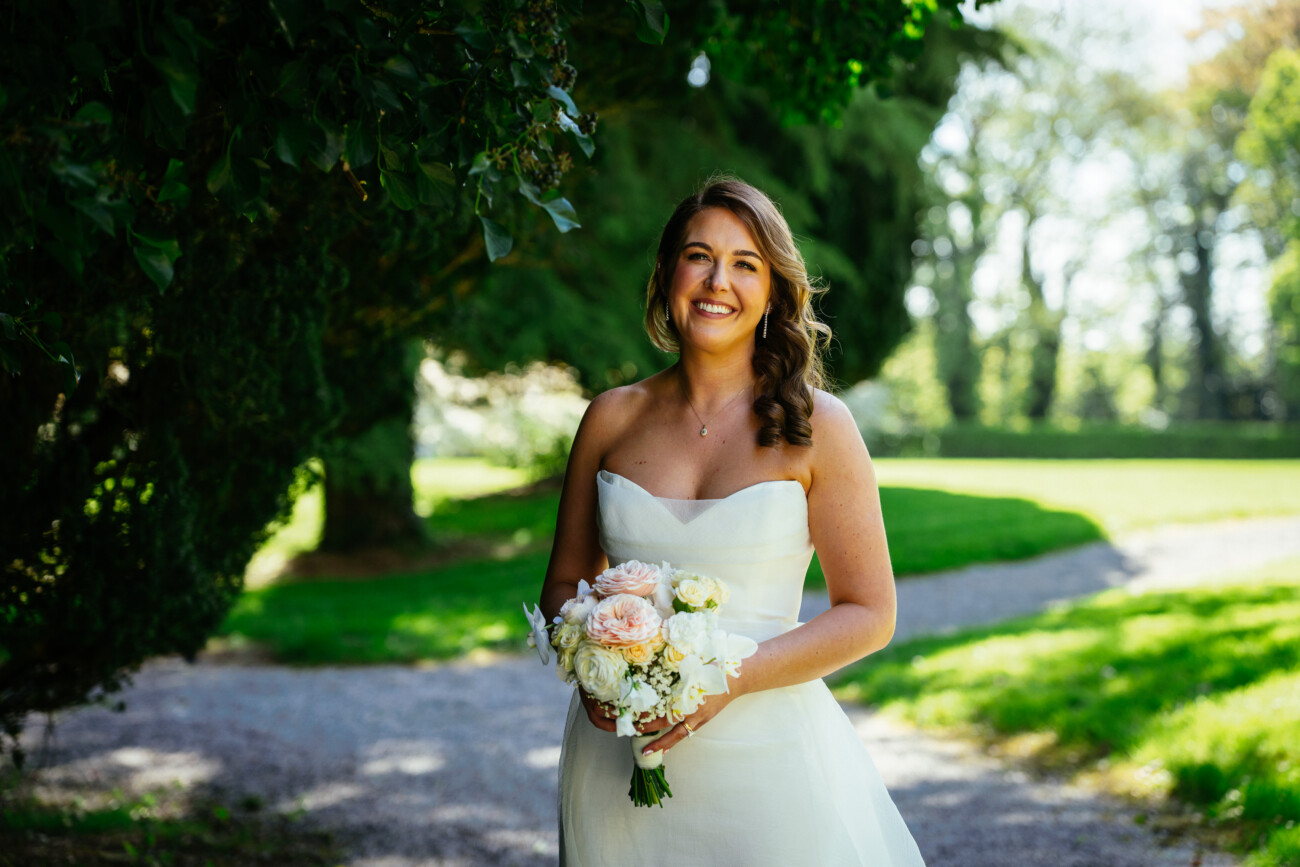 A woman in a white wedding dress holds a bouquet and smiles outdoors, with green trees and sunlight in the background, capturing the elegance of Castle Durrow Wedding Photography.