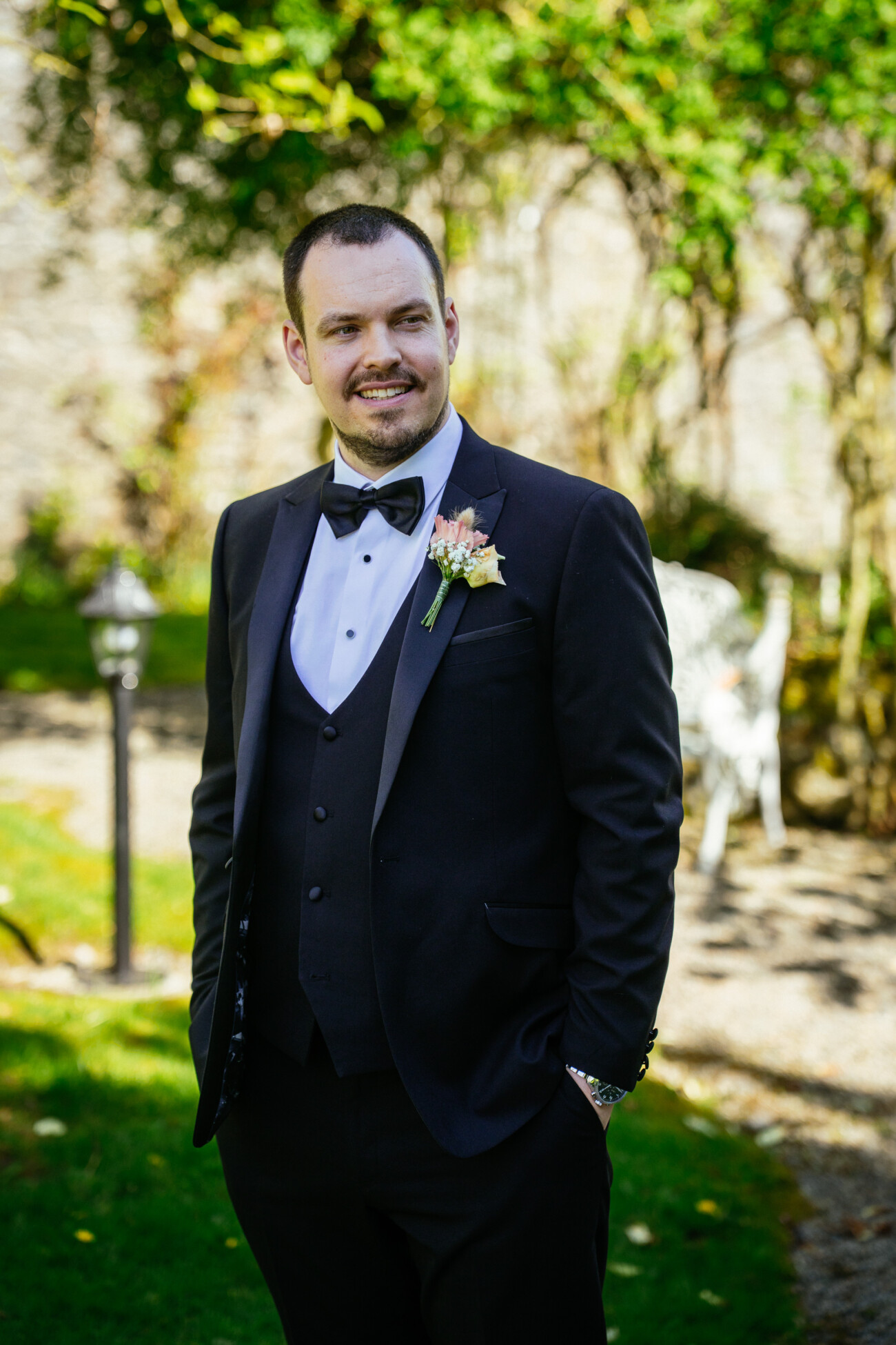 Man in a black dinner jacket with a buttonhole stands outdoors on grass, smiling, with trees and garden furniture in the background—capturing the timeless charm of Castle Durrow Wedding Photography.