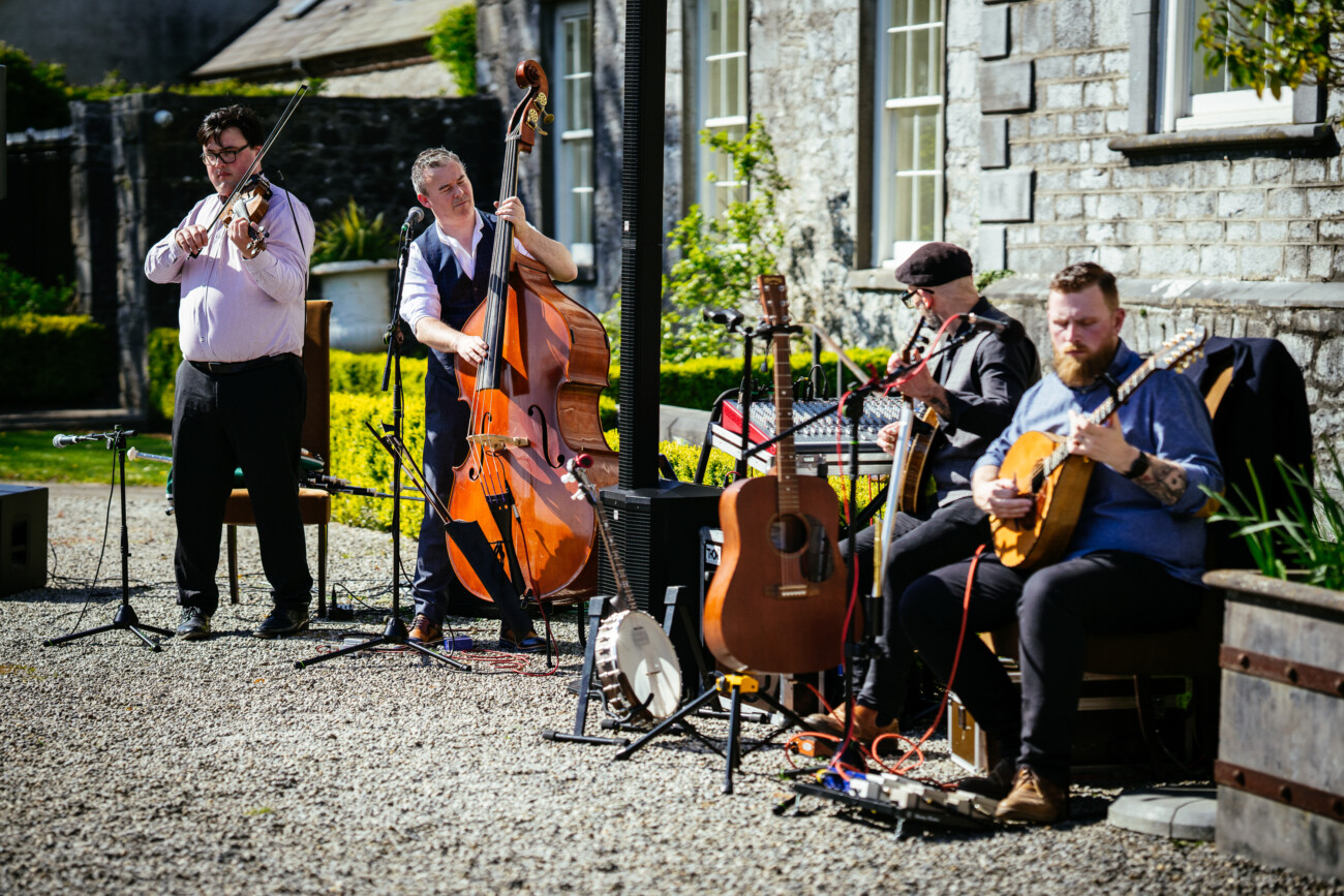 Four musicians play violin, double bass, accordion, and banjo outdoors near a stone building, their instruments and microphones set up around them on a sunny day. This lively scene captures the ambience often found in Castle Durrow wedding photography.