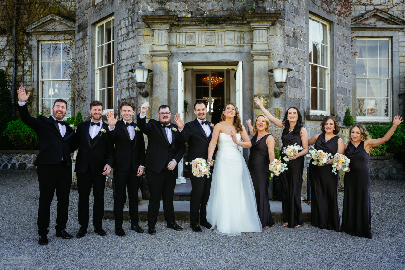 A bride and groom stand with their wedding party in formal attire, smiling and waving, in front of an old stone building, capturing a timeless moment of Castle Durrow Wedding Photography.