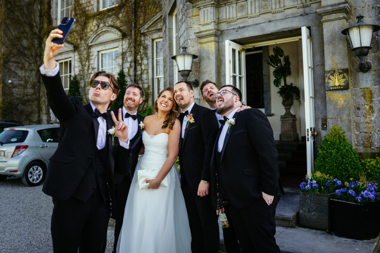 A bride and five men in formalwear pose for a selfie outside a stone building, smiling and making peace signs—a joyful moment beautifully captured in the style of Castle Durrow Wedding Photography.