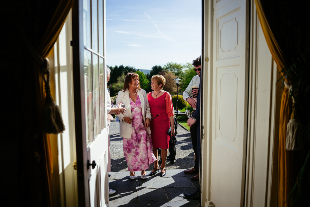 Two women in dresses stand outside an open doorway, conversing and smiling, with others nearby and greenery visible in the background—a perfect example of the candid moments captured in Castle Durrow Wedding Photography.
