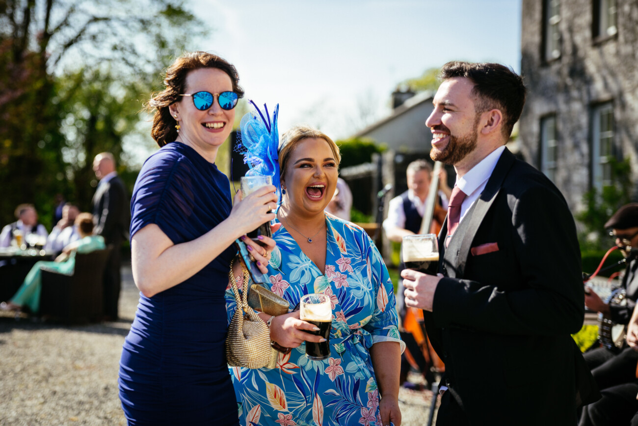 Three people stand outside at a social event, smiling and holding drinks, with other guests and the beautiful stone architecture of Castle Durrow visible in the background—capturing the joy and elegance characteristic of Castle Durrow wedding photography.