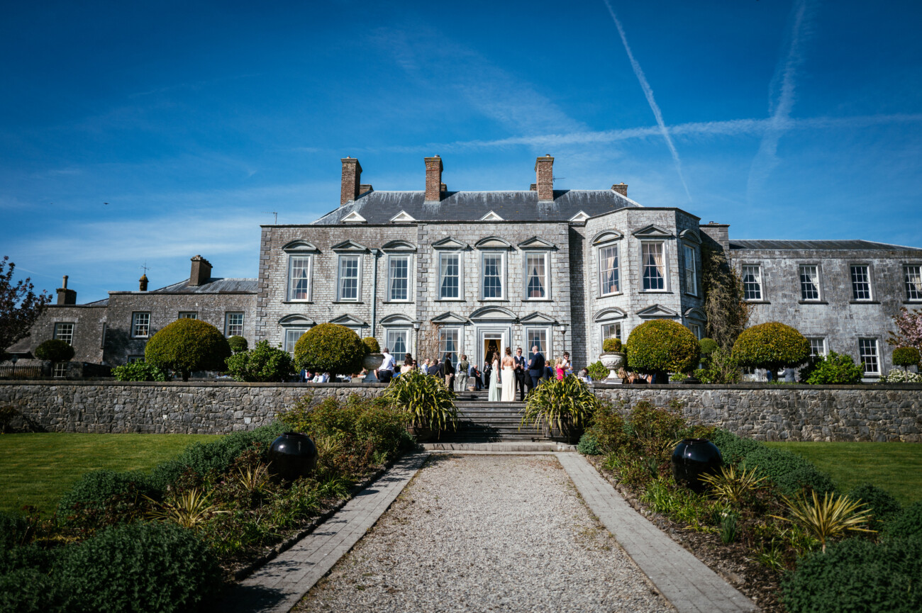 People are gathered outside a large, historic stone mansion on a sunny day, standing near the entrance with manicured gardens and a clear blue sky in the background—capturing the elegance and charm that defines Castle Durrow Wedding Photography.