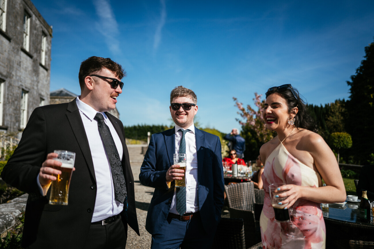 Three people in formal attire stand outdoors in sunlight, holding drinks and smiling, with a historic stone building and greenery in the background—a perfect moment captured through Castle Durrow Wedding Photography.
