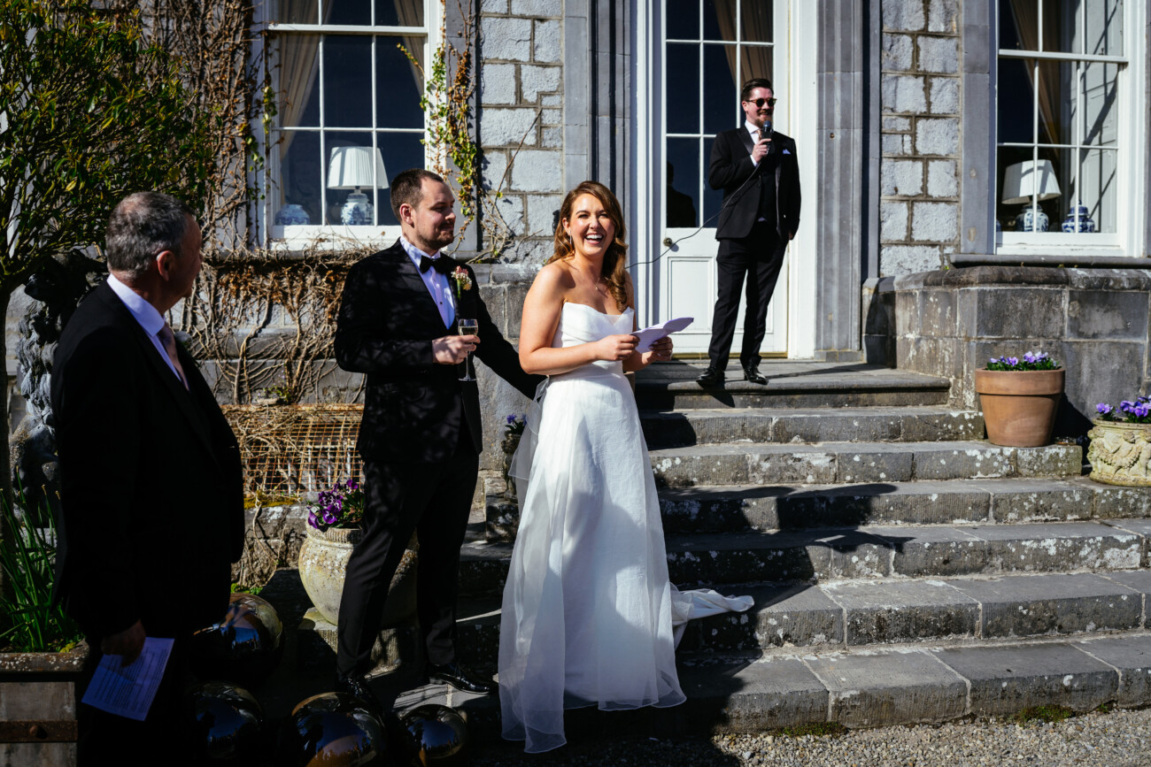 A bride in a white dress stands smiling with a groom and two men in suits on stone steps outside a building, holding papers and drinks—a timeless moment beautifully captured by Castle Durrow wedding photography.