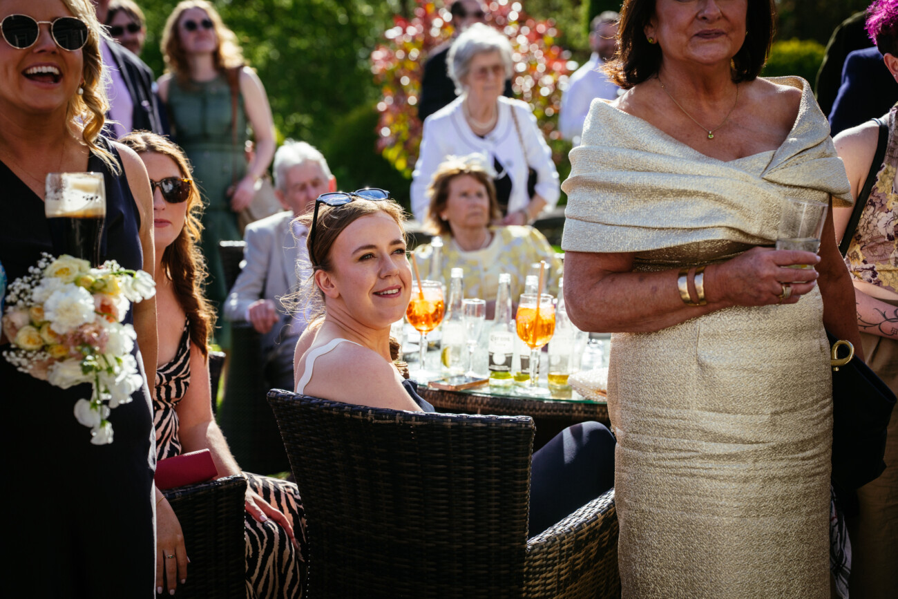 A group of people dressed formally gathers outdoors around a table with drinks, some standing and some seated, in bright sunlight—a perfect moment captured through Castle Durrow Wedding Photography.