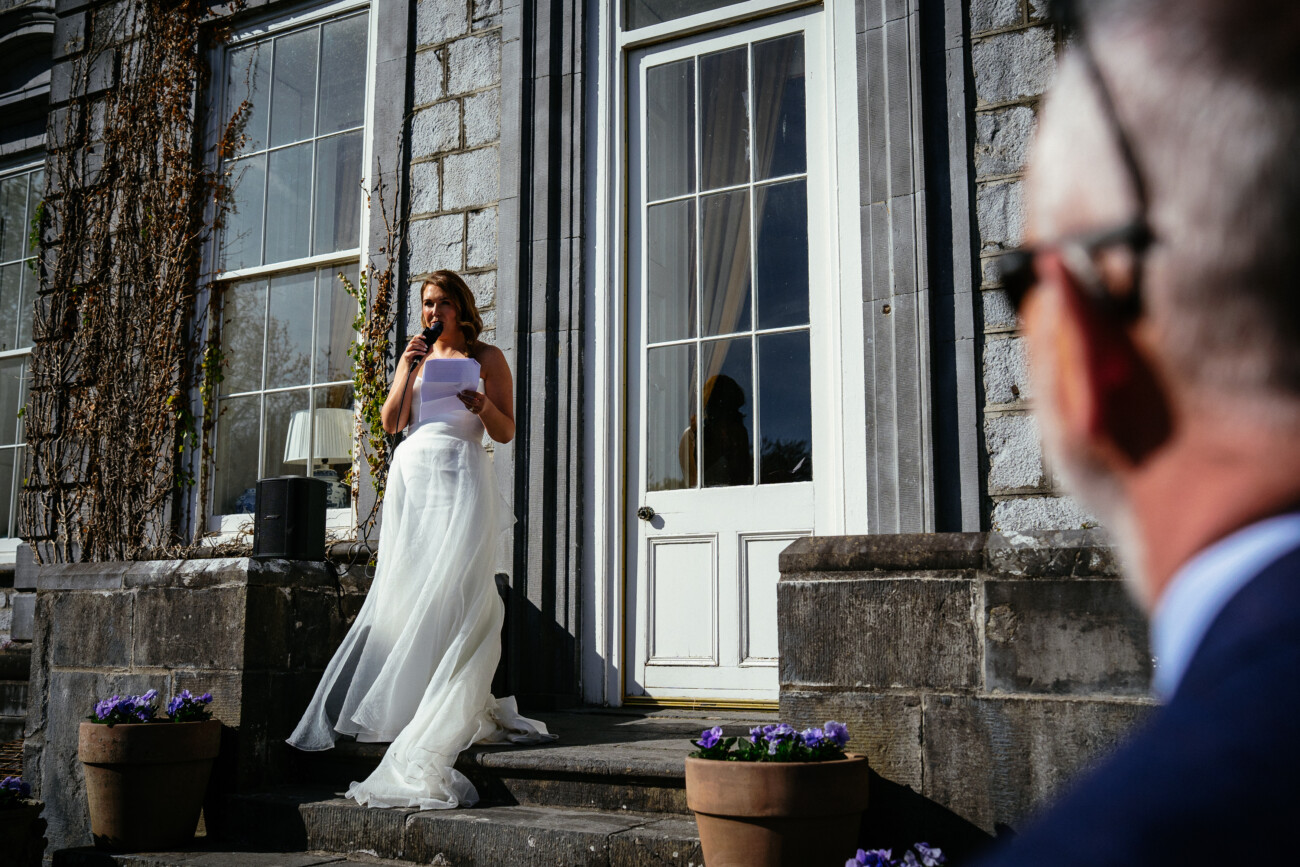 A bride in a white dress stands on the stone steps of Castle Durrow, holding a microphone and paper as she speaks, while a man in sunglasses looks on in the foreground—an elegant moment captured through Castle Durrow wedding photography.