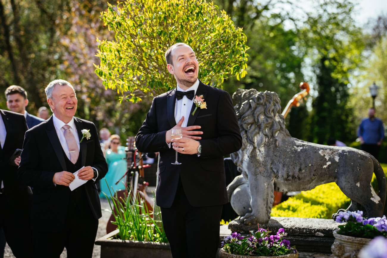 Two men in suits laugh outdoors near a stone lion statue and garden, one holding a drink, during a sunny event—a perfect moment captured by Castle Durrow Wedding Photography.