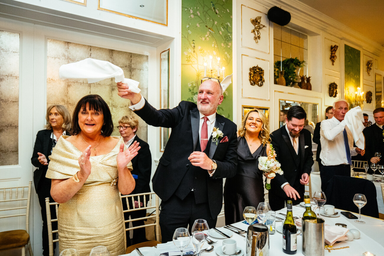 A group of well-dressed people stand and wave serviettes in a decorated dining room, appearing to celebrate at a formal event or reception, perfectly capturing the joyful atmosphere that Castle Durrow Wedding Photography is known for.