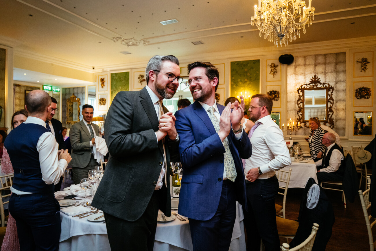 Two men in suits stand and smile at a formal dinner event, surrounded by other guests who are seated or standing in an elegant, well-lit dining room—capturing the refined atmosphere often seen in Castle Durrow Wedding Photography.