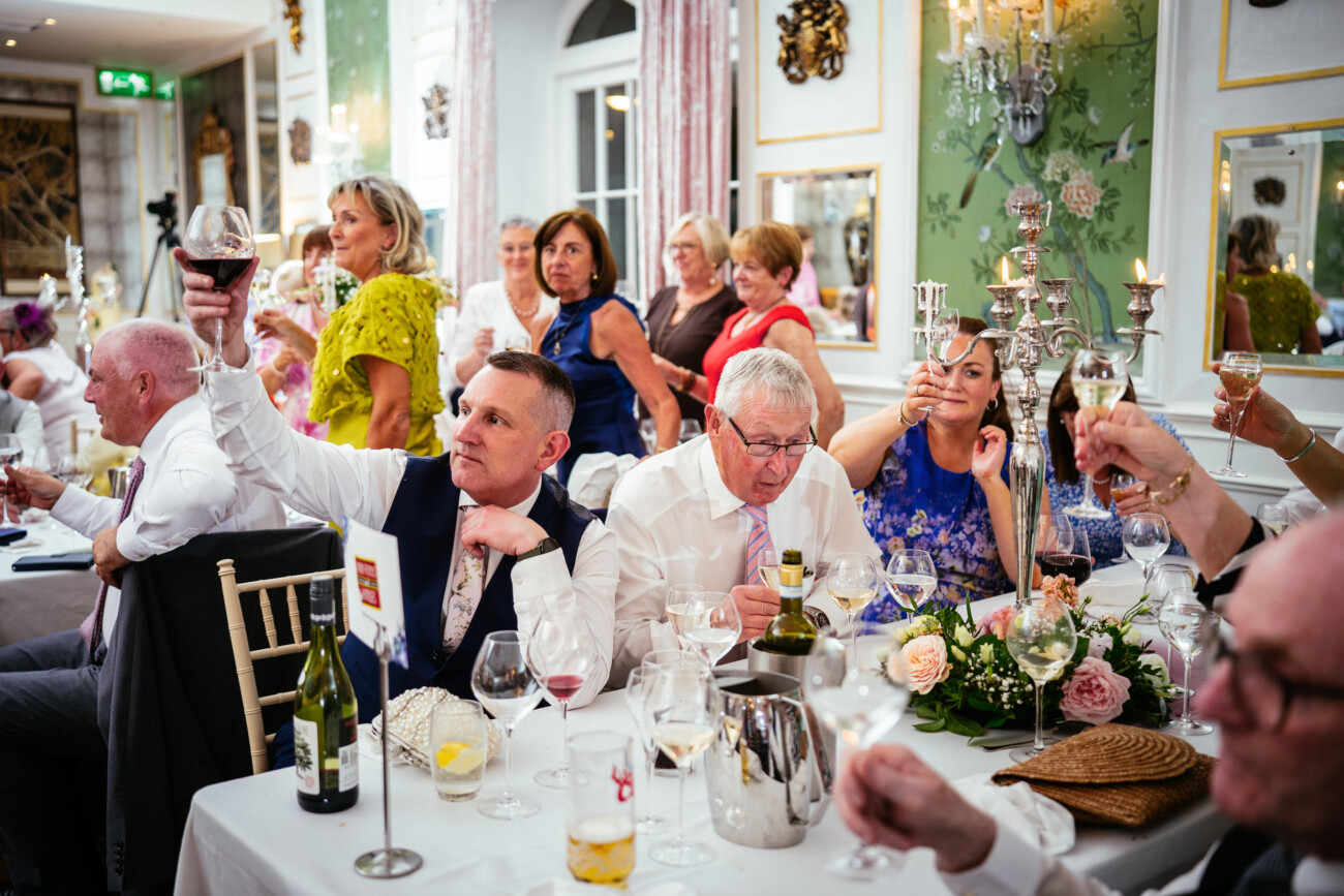 Guests seated at decorated tables raise glasses in a toast at a formal indoor event, with floral arrangements and candles in the background—a perfect moment captured by Castle Durrow Wedding Photography.