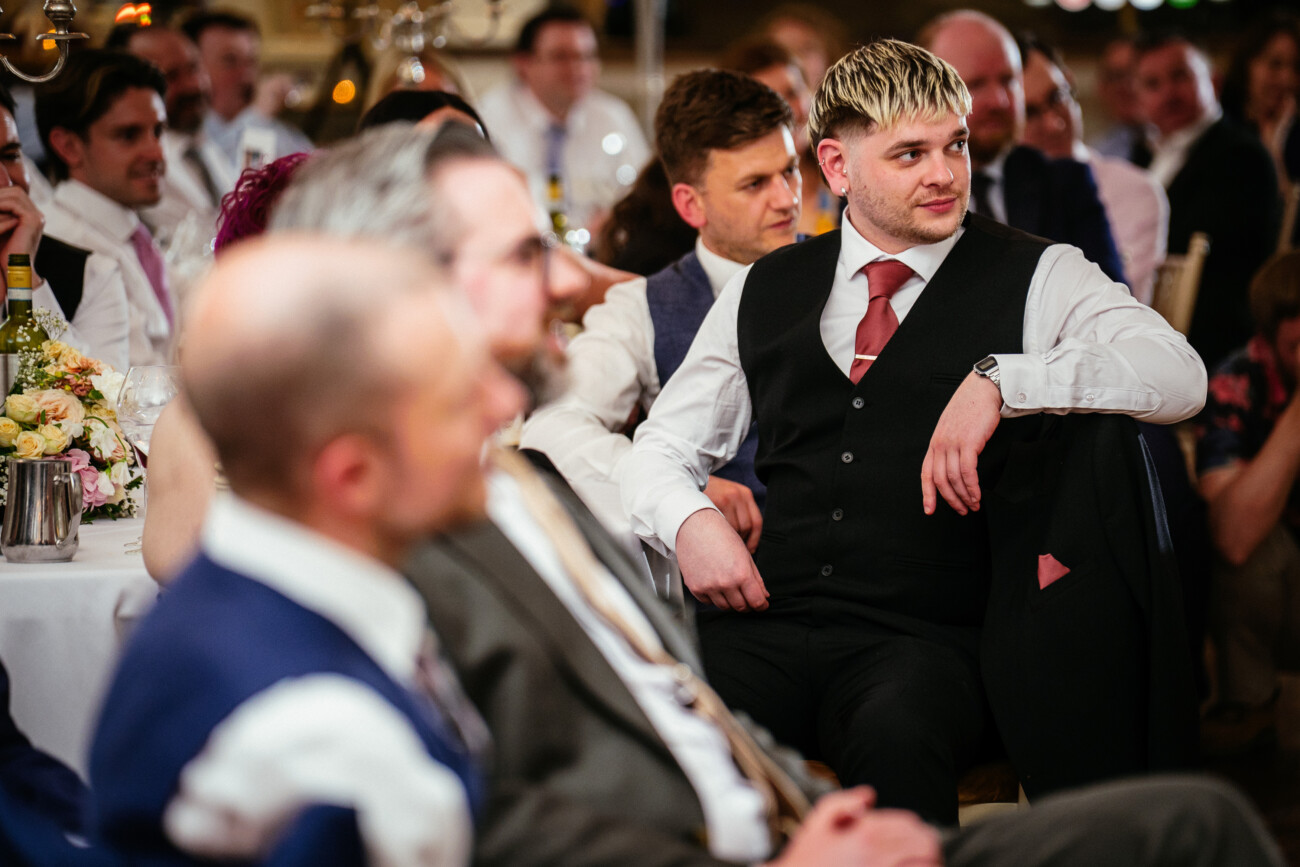 A group of people dressed formally sit at tables during an indoor event, listening attentively. One man in a black waistcoat and red tie sits in focus among the group, capturing the elegant ambience typical of Castle Durrow Wedding Photography.