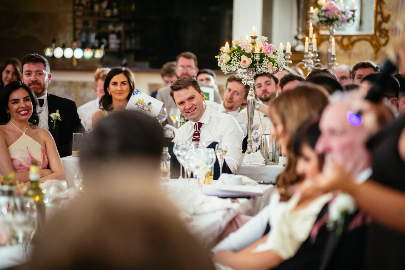 A group of people at a formal event sit around decorated tables, listening attentively, with candles, flowers, and drinks visible in the scene—an elegant atmosphere beautifully captured in classic Castle Durrow Wedding Photography.