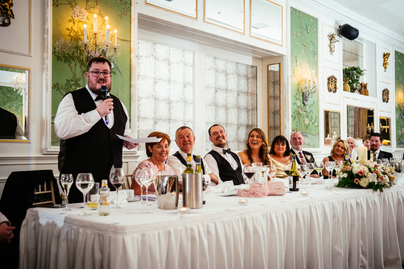 A man stands giving a speech with a microphone at a formal banquet table, whilst seven people seated beside him listen and smile in an elegant, decorated room—capturing the timeless charm of Castle Durrow Wedding Photography.