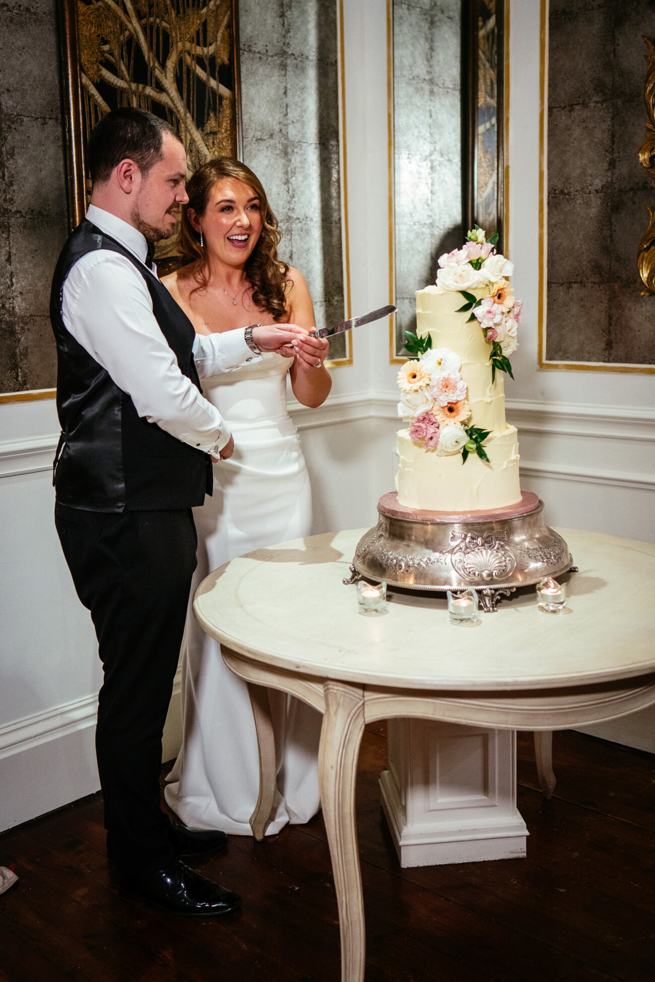 A bride and groom stand together cutting a tall wedding cake decorated with flowers, placed on a round table in an elegant room, beautifully captured by Castle Durrow Wedding Photography.