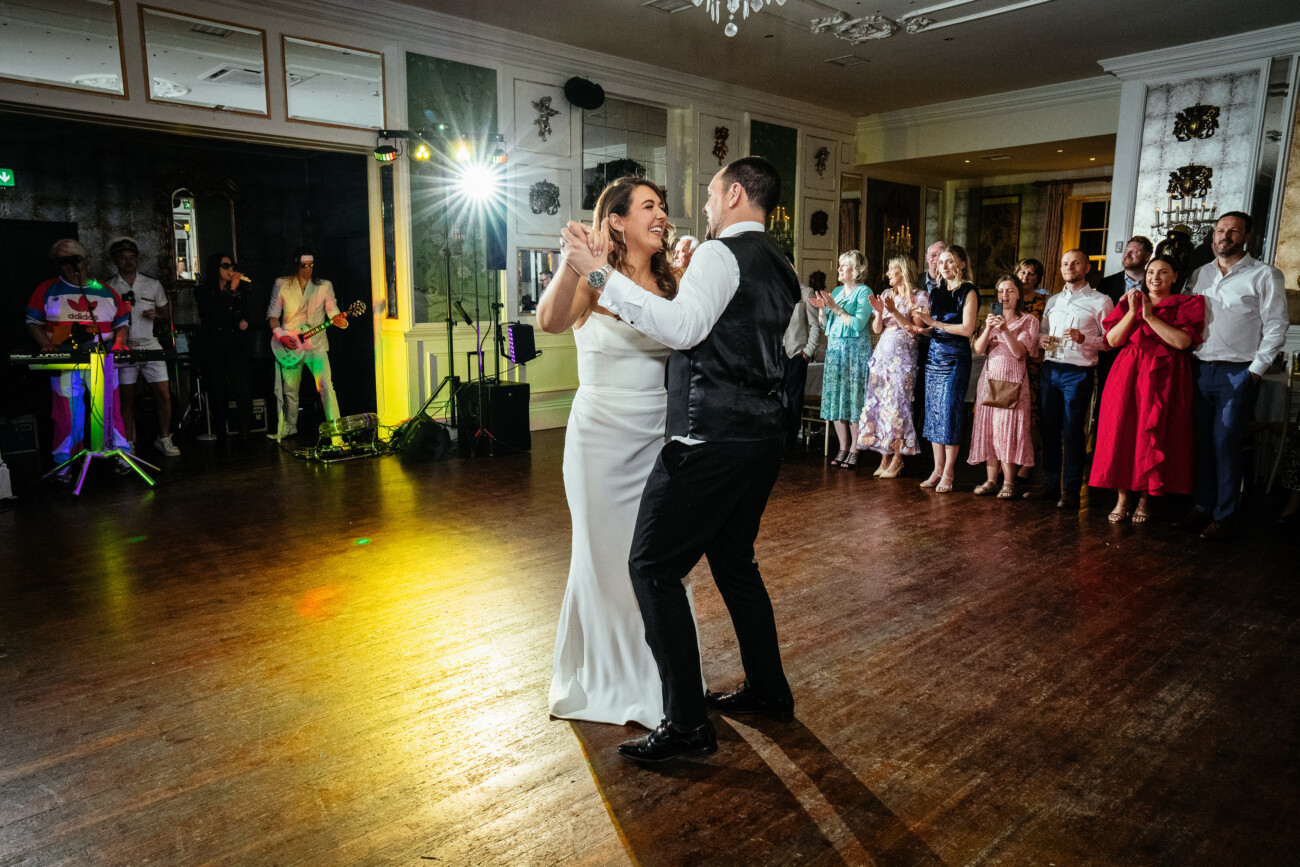 A bride and groom dance together on a wooden floor while guests watch and a live band plays in the background at a wedding reception, capturing the elegance and romance that define Castle Durrow Wedding Photography.