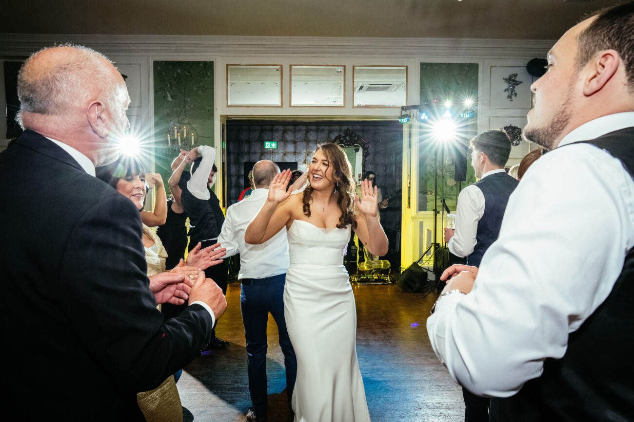 A bride in a white dress dances and smiles with guests on a lively, well-lit dance floor at an indoor wedding reception, beautifully captured in classic Castle Durrow Wedding Photography style.