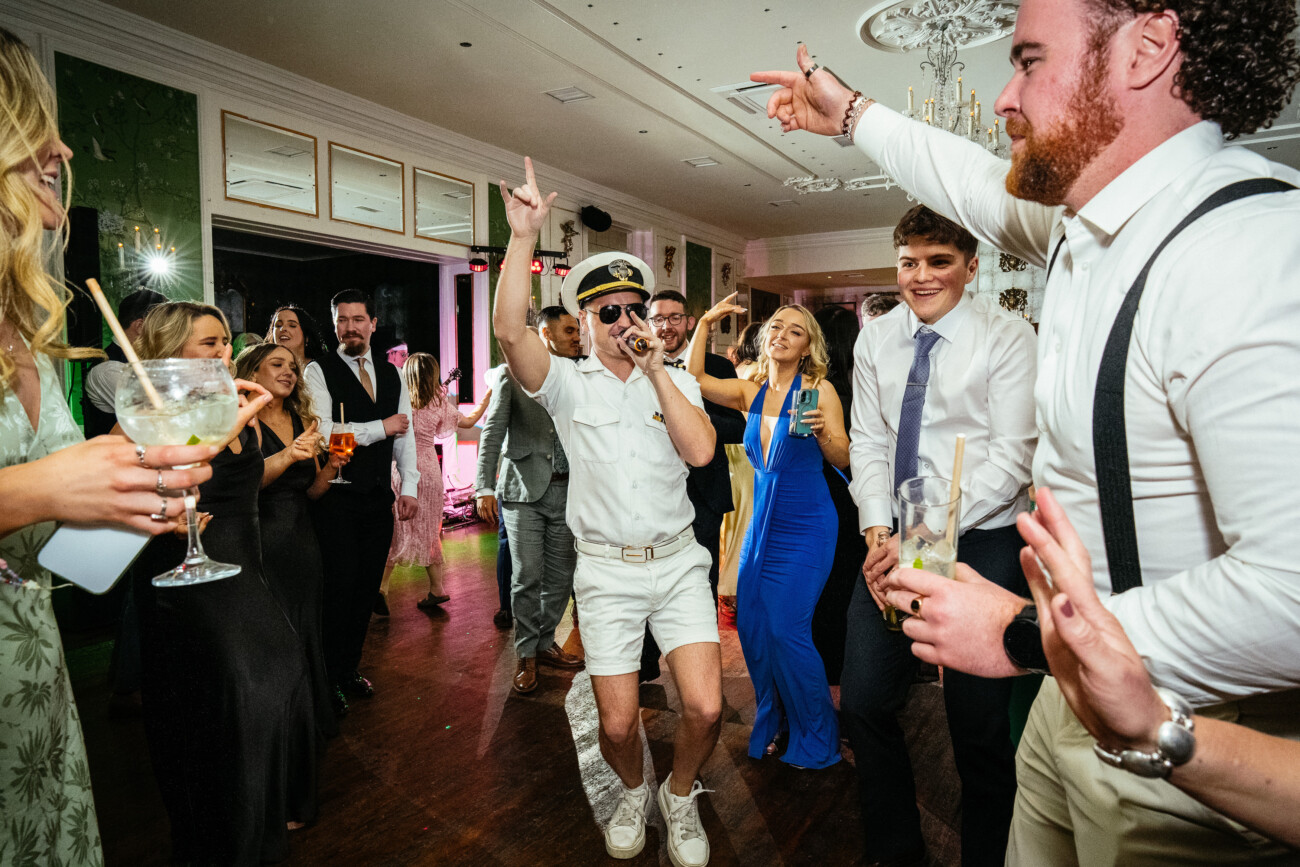 A man dressed as a captain sings into a microphone as people in formal attire dance and celebrate at a lively indoor party, perfectly capturing the vibrant atmosphere often seen in Castle Durrow Wedding Photography.