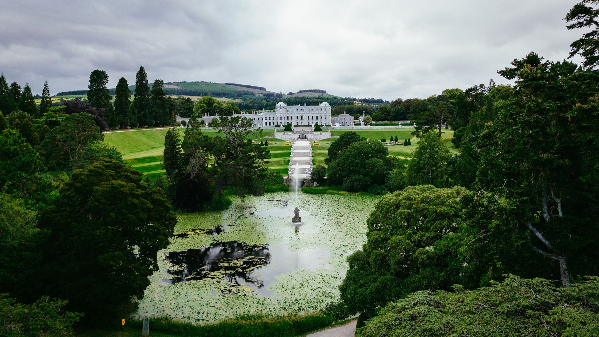 Powerscourt Estate Wicklow Ireland seen from above, with a fountain in a pond, surrounded by green lawns, trees, and distant hills under a cloudy sky—an idyllic setting worthy of inclusion among the Top 20 Wedding Venues in Ireland 2026.