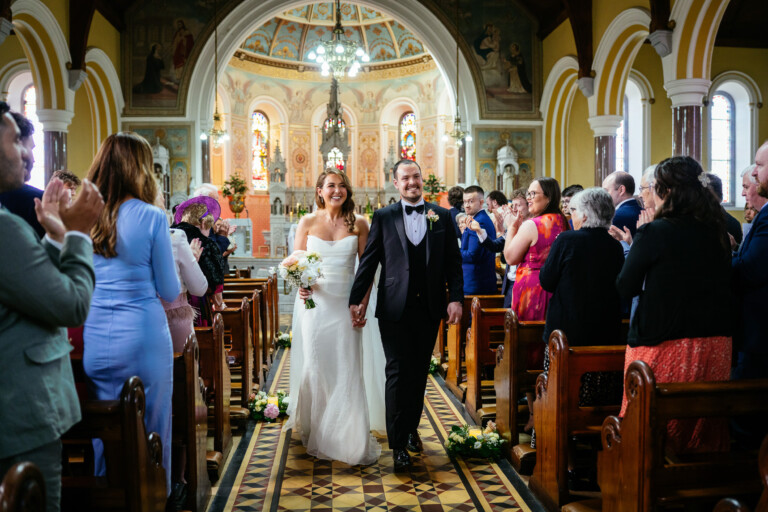 A Cherry Blossom Wedding at Castle Durrow: Lauren & David 126 A bride and groom walk down the aisle in a church, smiling and holding hands, as guests stand and applaud on either side, capturing a timeless moment reminiscent of Castle Durrow Wedding Photography.