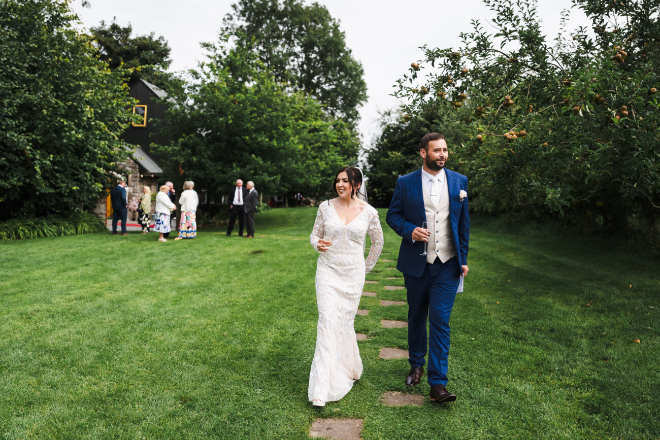 A Relaxed Summer Wedding at Village Barn, Tyrellspass | Amy & Eoghan 68 A bride in a white dress and a groom in a blue suit walk on a garden path, holding drinks, with guests and trees in the background.