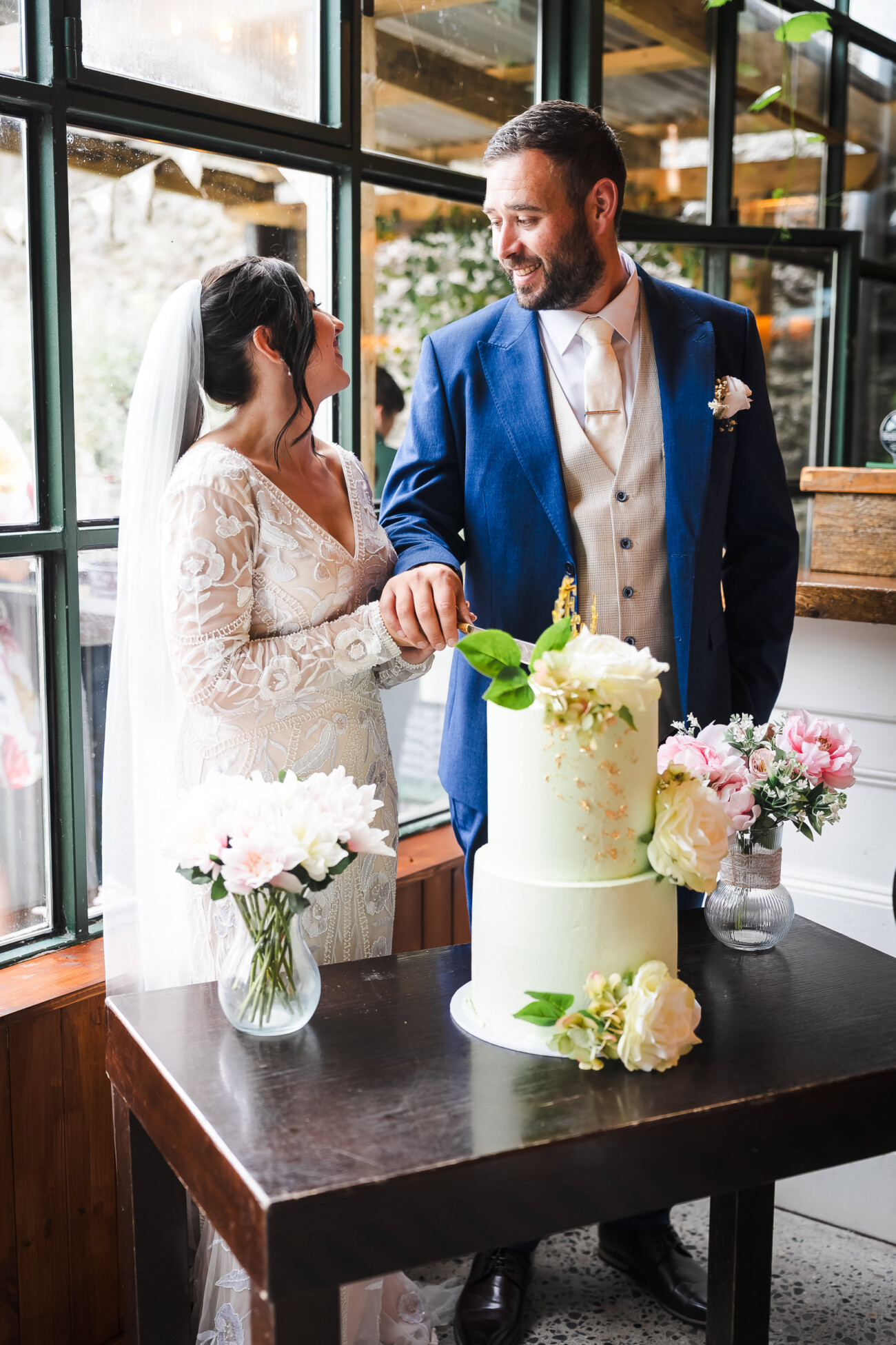 A Relaxed Summer Wedding at Village Barn, Tyrellspass | Amy & Eoghan 69 A bride and groom stand together beside a white wedding cake decorated with flowers, holding hands and smiling at one another in a bright indoor setting.