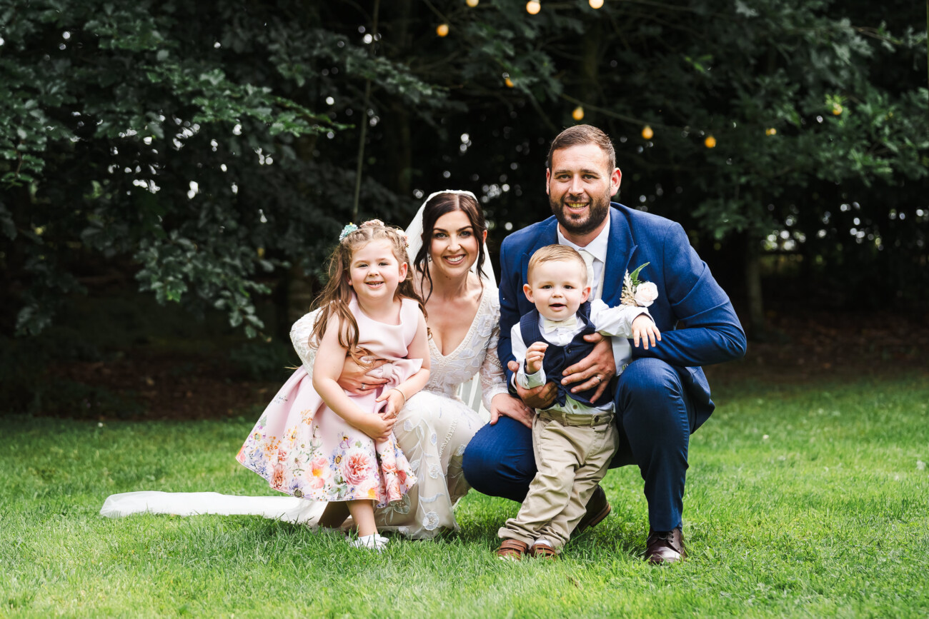A Relaxed Summer Wedding at Village Barn, Tyrellspass | Amy & Eoghan 70 A bride and groom kneel on grass outdoors with a young girl in a floral dress and a toddler boy, all smiling at the camera. Trees and fairy lights are in the background.