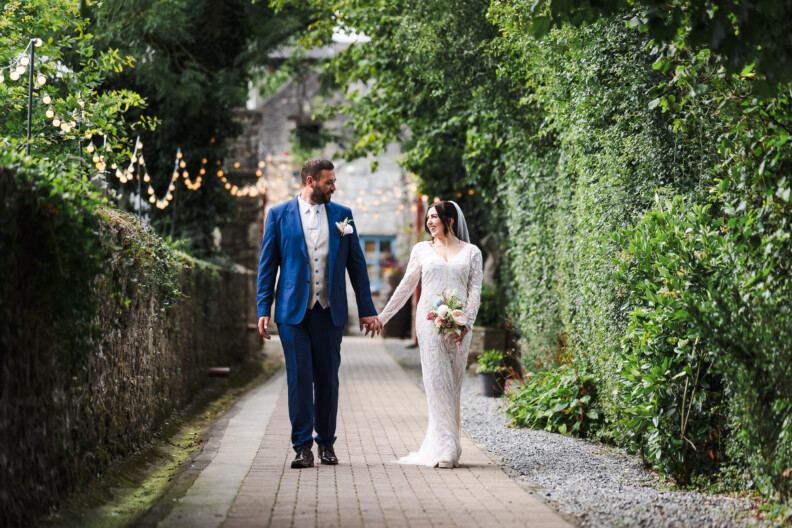 A bride and groom walk hand in hand down a garden path lined with greenery and fairy lights, smiling at each other.