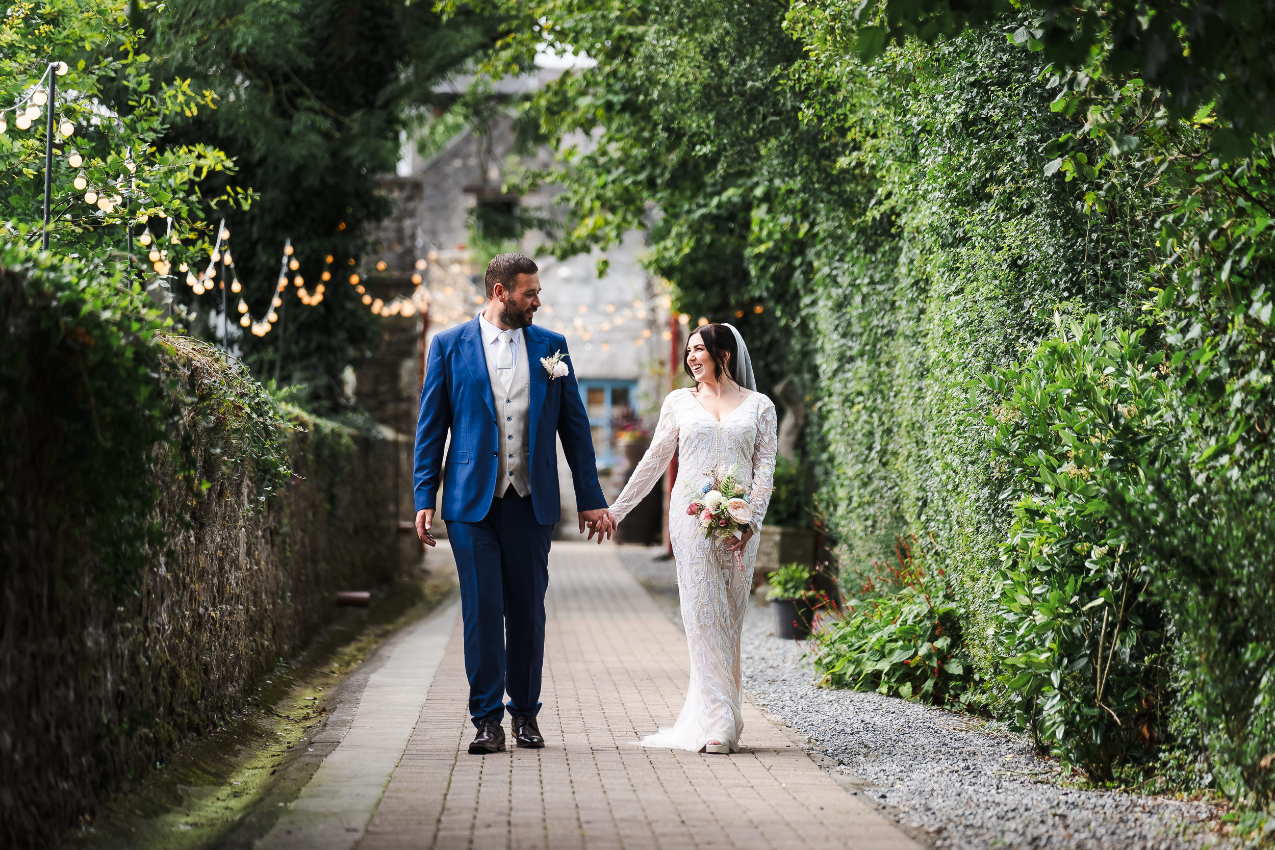 A bride and groom walk hand in hand down a garden path lined with greenery and fairy lights, smiling at each other.