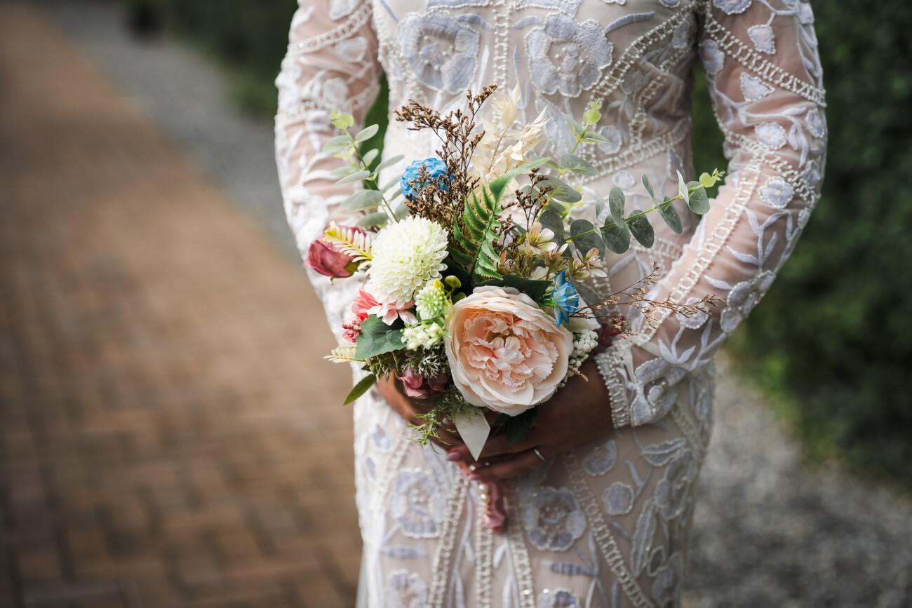 A Relaxed Summer Wedding at Village Barn, Tyrellspass | Amy & Eoghan 72 Person in a detailed white lace wedding dress holding a bouquet of mixed flowers, including roses, greenery, and chrysanthemums, standing on a brick path outdoors.