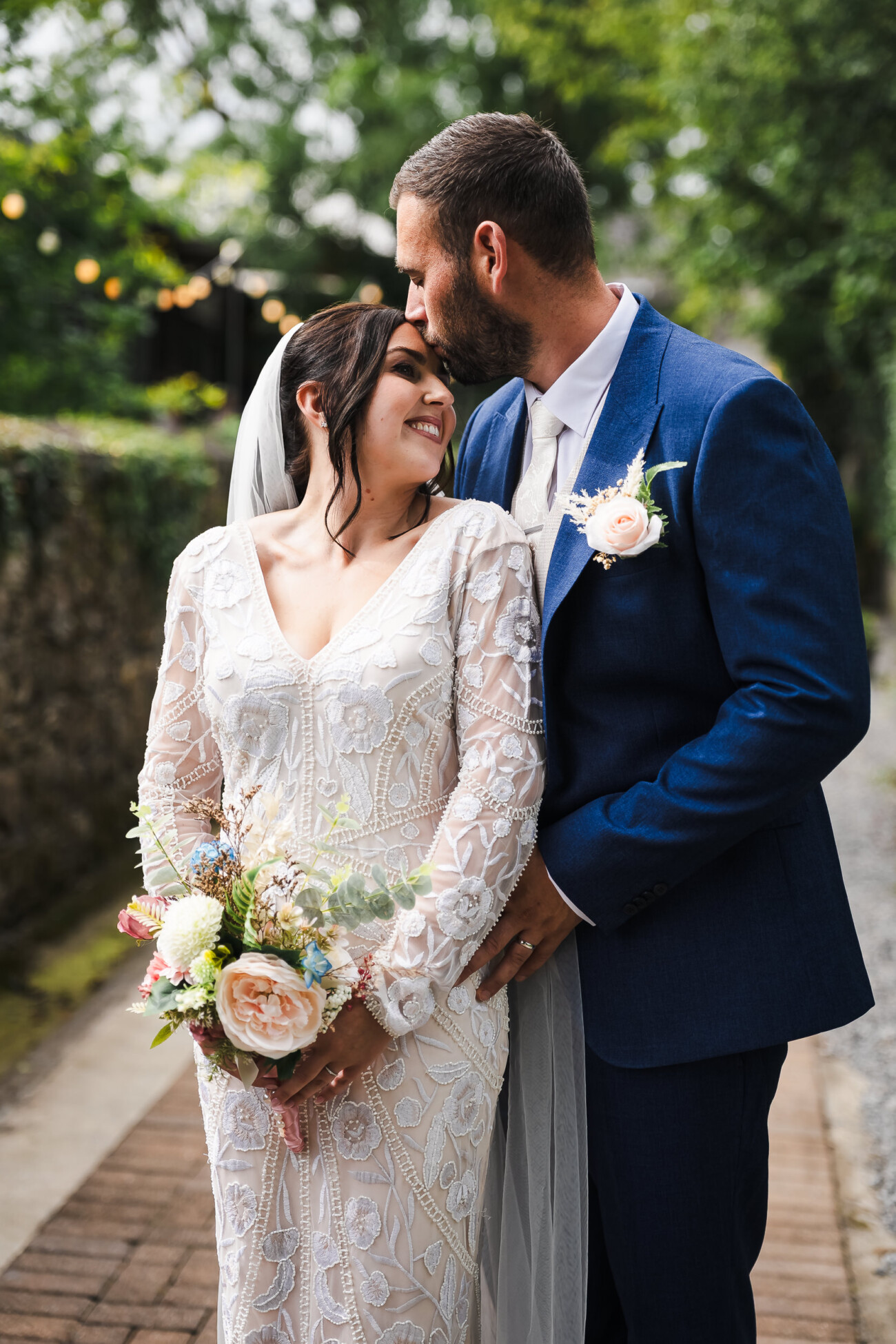 A Relaxed Summer Wedding at Village Barn, Tyrellspass | Amy & Eoghan 73 A bride in a lace dress holds a bouquet and smiles as the groom in a blue suit kisses her head outdoors on a pathway.