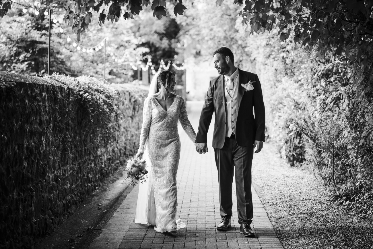 A Relaxed Summer Wedding at Village Barn, Tyrellspass | Amy & Eoghan 74 A bride and groom walk hand in hand down a tree-lined path, dressed in formal wedding attire, looking at each other and smiling.
