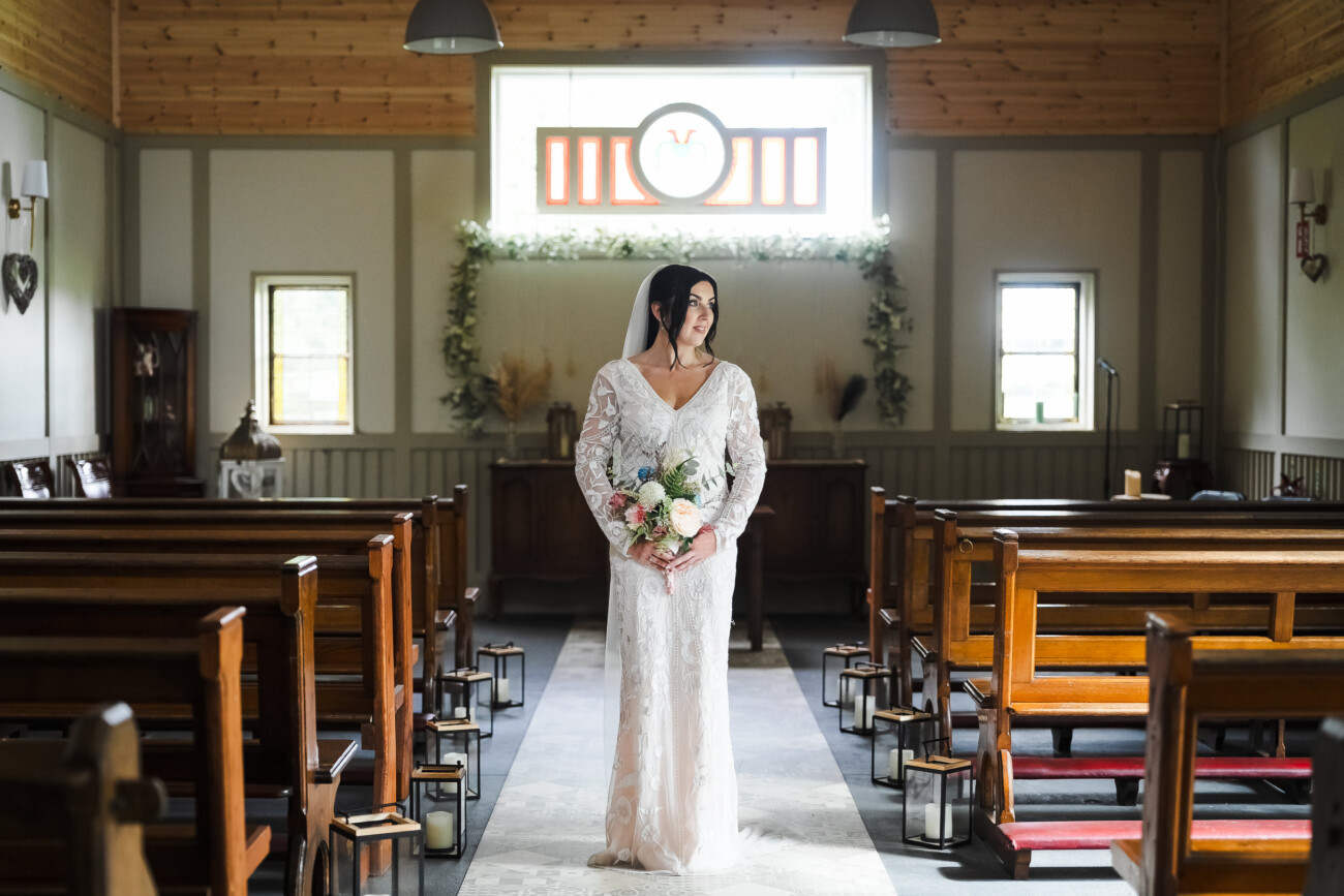 A Relaxed Summer Wedding at Village Barn, Tyrellspass | Amy & Eoghan 75 A bride in a white lace dress stands holding a bouquet in the aisle of a small, sunlit chapel with wooden pews and a stained glass window behind her.