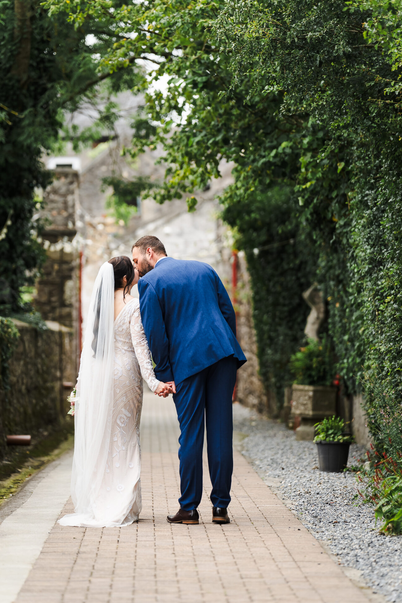 A Relaxed Summer Wedding at Village Barn, Tyrellspass | Amy & Eoghan 76 A bride and groom stand on a tree-lined path, holding hands and sharing a kiss, with greenery and stone walls around them.