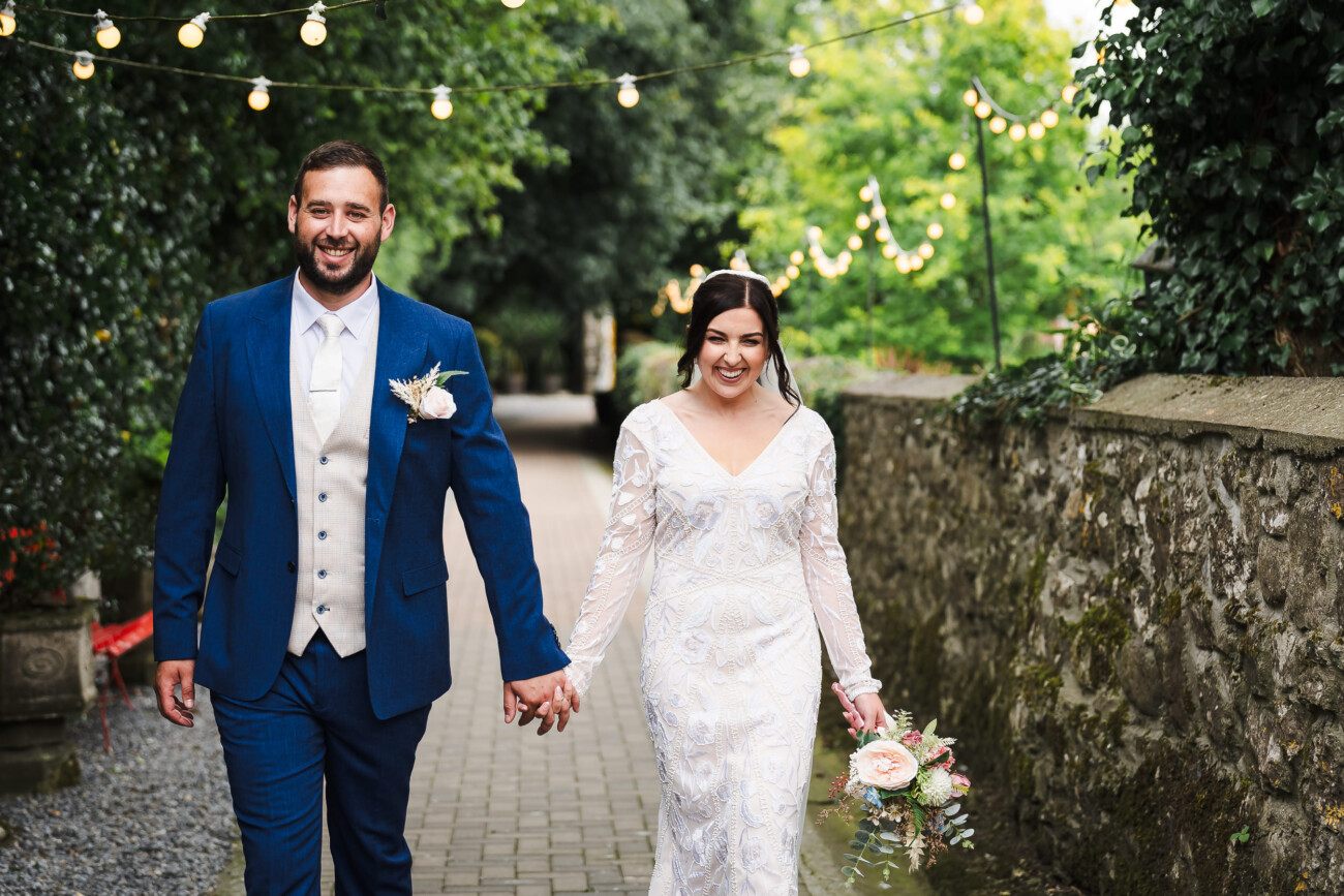 A Relaxed Summer Wedding at Village Barn, Tyrellspass | Amy & Eoghan 77 A bride and groom walk hand in hand outdoors, both smiling. The bride holds a bouquet, and fairy lights hang above them along a path bordered by greenery and a stone wall.