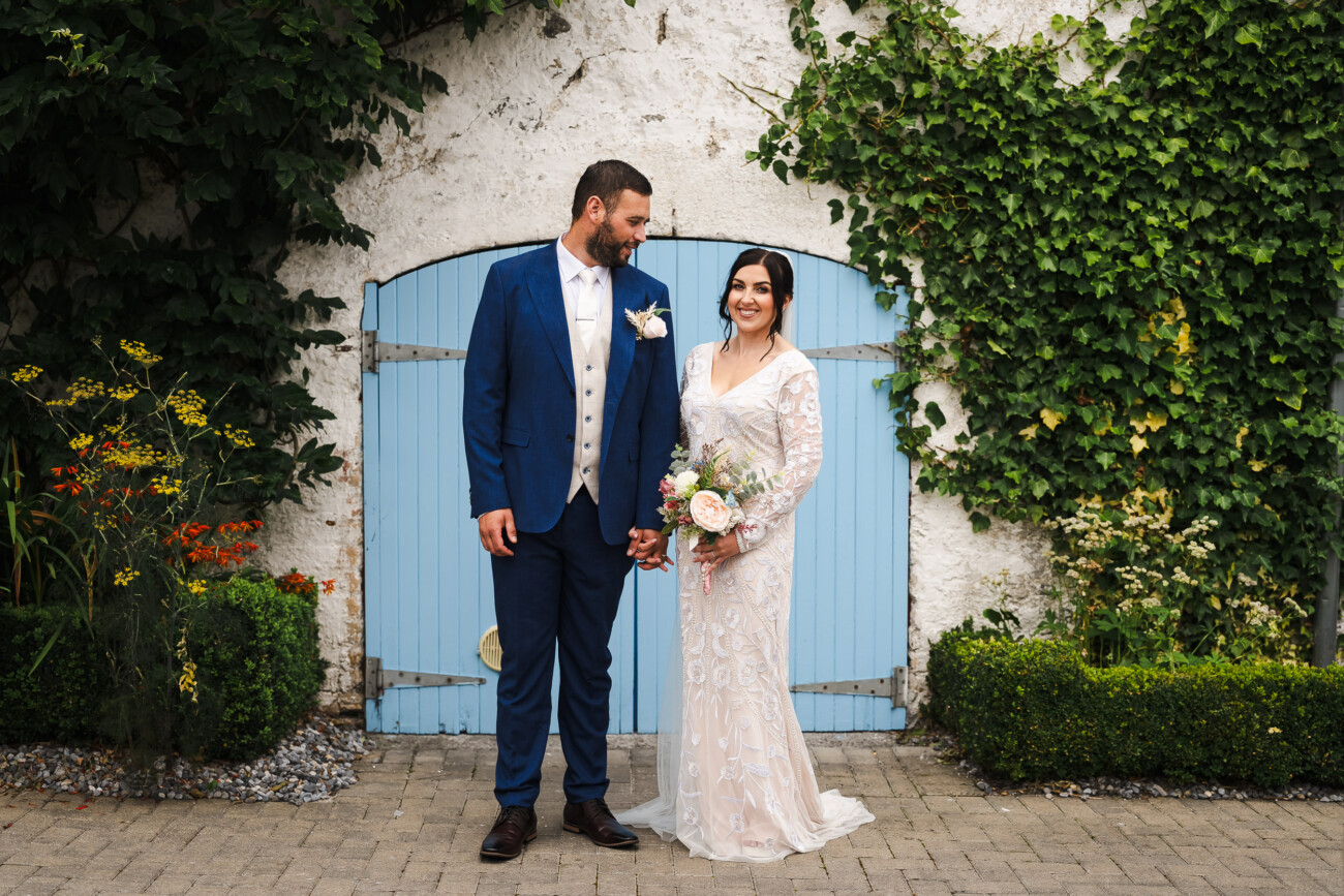 A Relaxed Summer Wedding at Village Barn, Tyrellspass | Amy & Eoghan 78 A bride in a white dress and a groom in a blue suit stand holding hands in front of blue doors, surrounded by greenery.