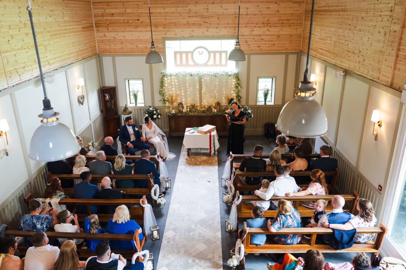 A Relaxed Summer Wedding at Village Barn, Tyrellspass | Amy & Eoghan 33 A wedding ceremony takes place in a small, decorated chapel with guests seated in wooden pews and an officiant standing at the front.