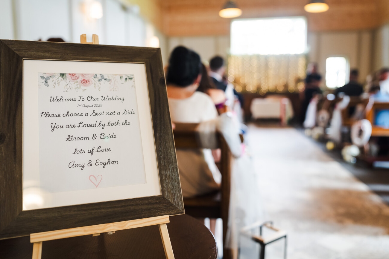 A Relaxed Summer Wedding at Village Barn, Tyrellspass | Amy & Eoghan 34 A framed wedding welcome sign on an easel is in focus, with seated guests and an officiant blurred in the background inside a decorated venue.