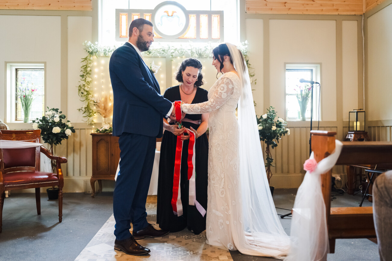 A Relaxed Summer Wedding at Village Barn, Tyrellspass | Amy & Eoghan 35 A couple stands facing each other during a wedding ceremony as an officiant ties red and white ribbons around their hands inside a decorated room.