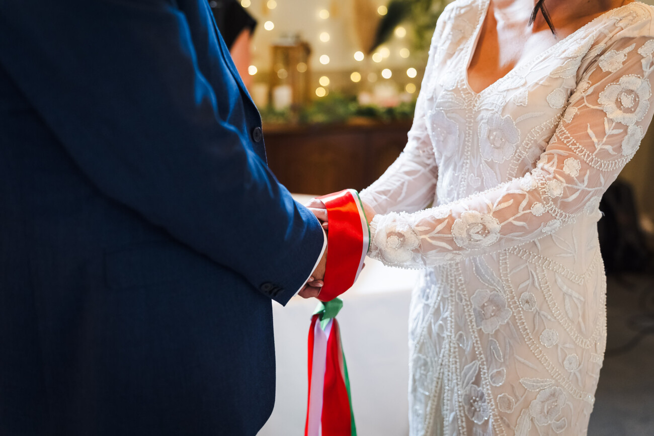 A Relaxed Summer Wedding at Village Barn, Tyrellspass | Amy & Eoghan 36 Two people stand facing each other, holding hands wrapped together with a red and green ribbon, one wearing a white dress and the other a blue suit.