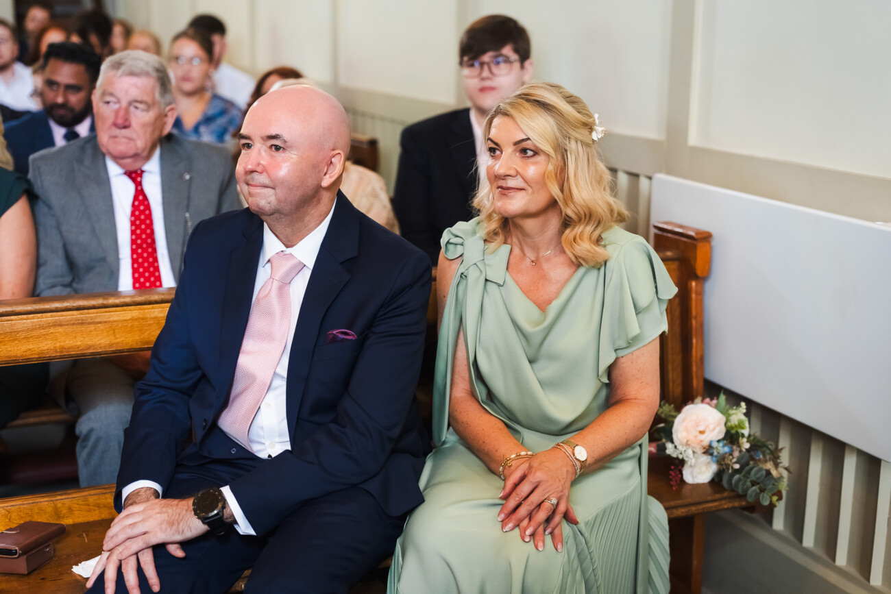 A Relaxed Summer Wedding at Village Barn, Tyrellspass | Amy & Eoghan 37 A man in a suit and a woman in a green dress sit together on a wooden pew in a room with other seated people, some dressed formally. Flowers rest on the pew beside them.