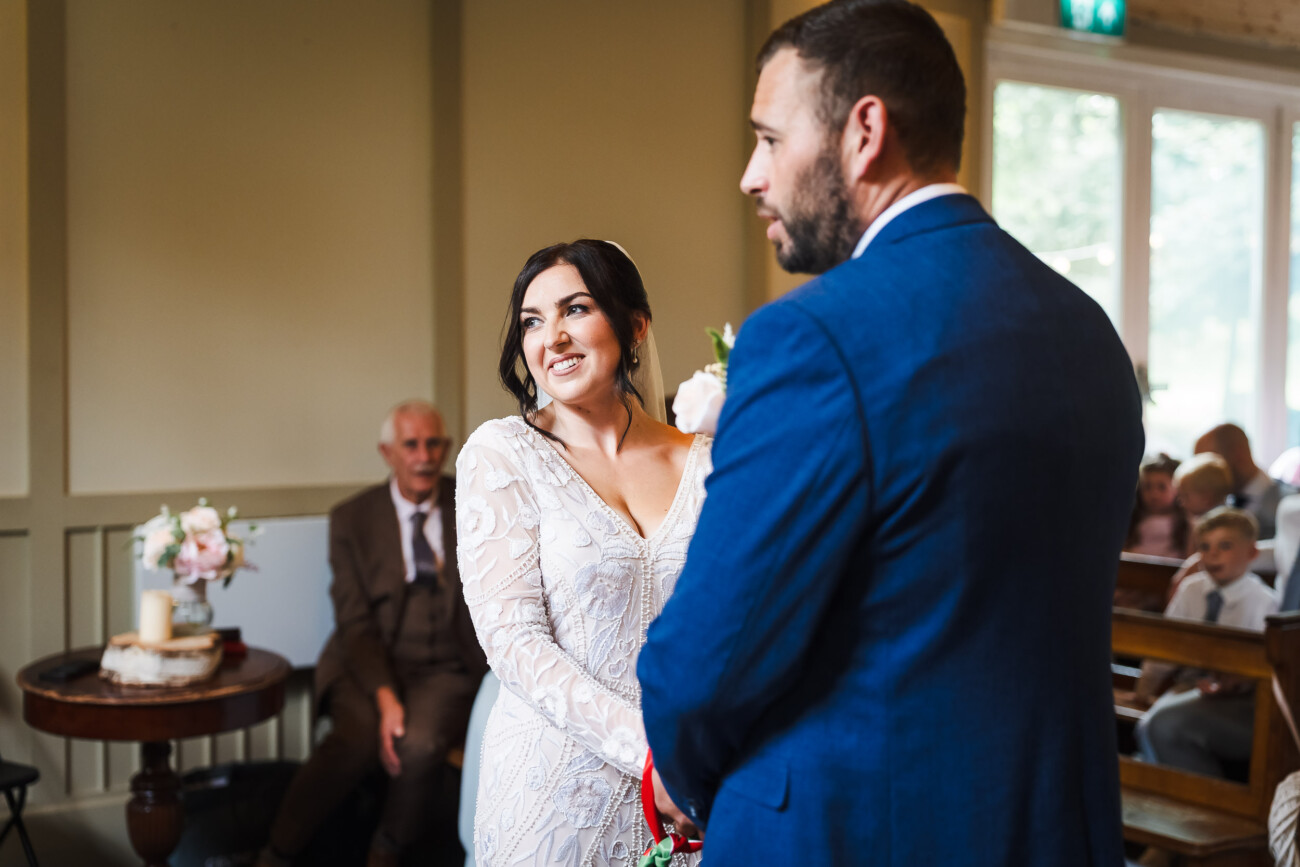 A Relaxed Summer Wedding at Village Barn, Tyrellspass | Amy & Eoghan 38 A bride in a white dress stands beside a man in a blue suit during a wedding ceremony, with guests seated in the background.