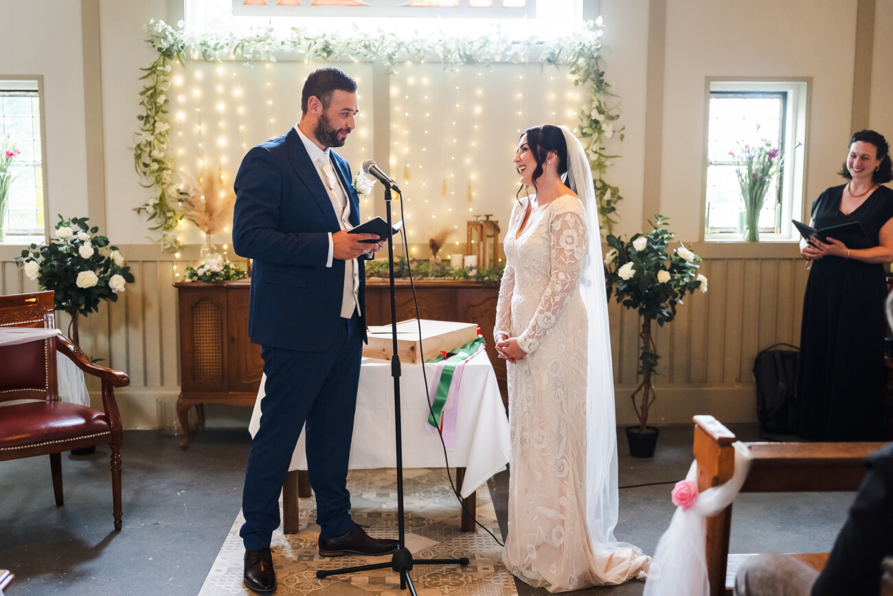 A Relaxed Summer Wedding at Village Barn, Tyrellspass | Amy & Eoghan 39 A groom in a blue suit reads vows to a bride in a white lace dress during their wedding ceremony inside a decorated room with floral arrangements.