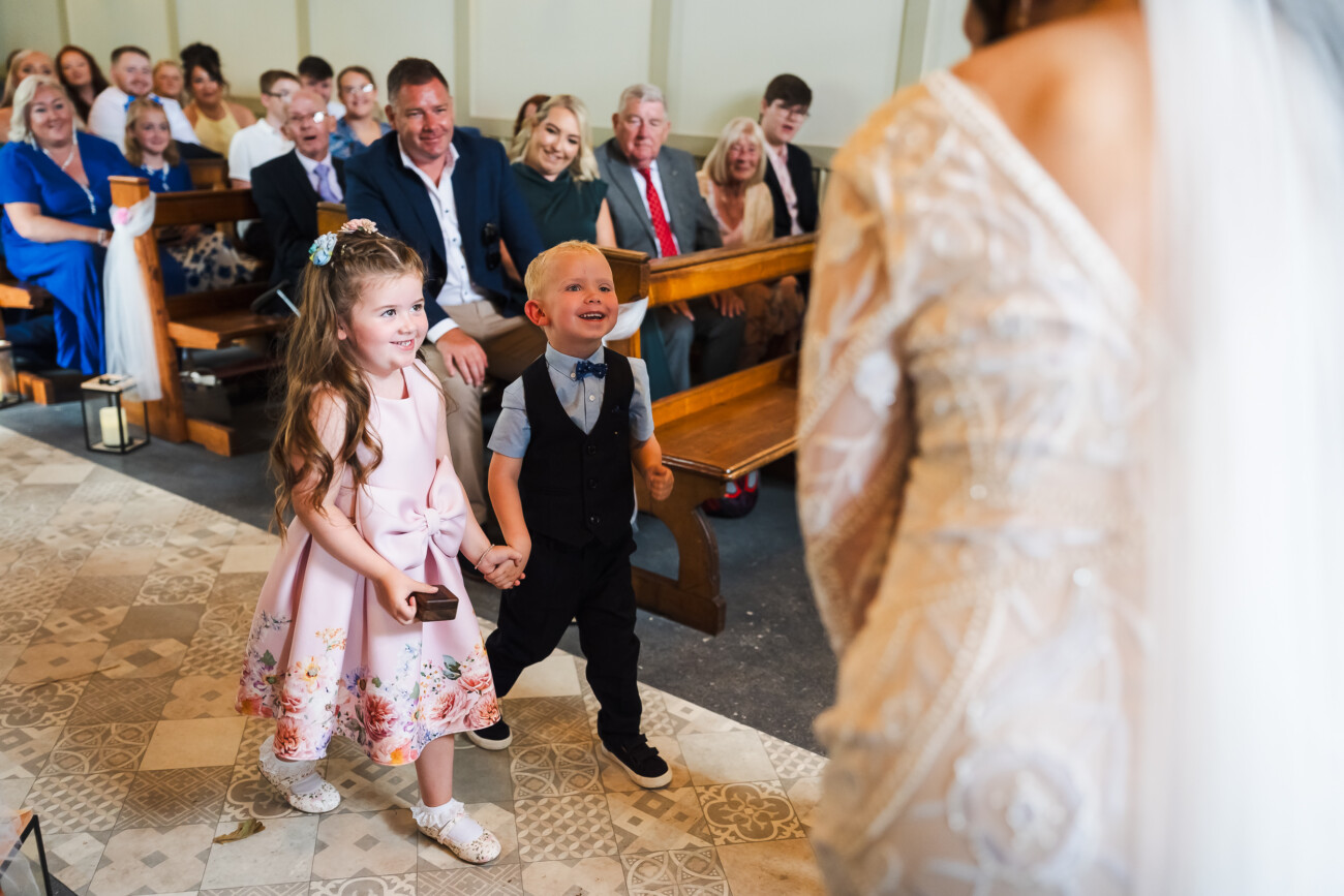 A Relaxed Summer Wedding at Village Barn, Tyrellspass | Amy & Eoghan 40 Two young children walk up the aisle holding hands, smiling at a bride during a wedding ceremony as guests watch from wooden pews.
