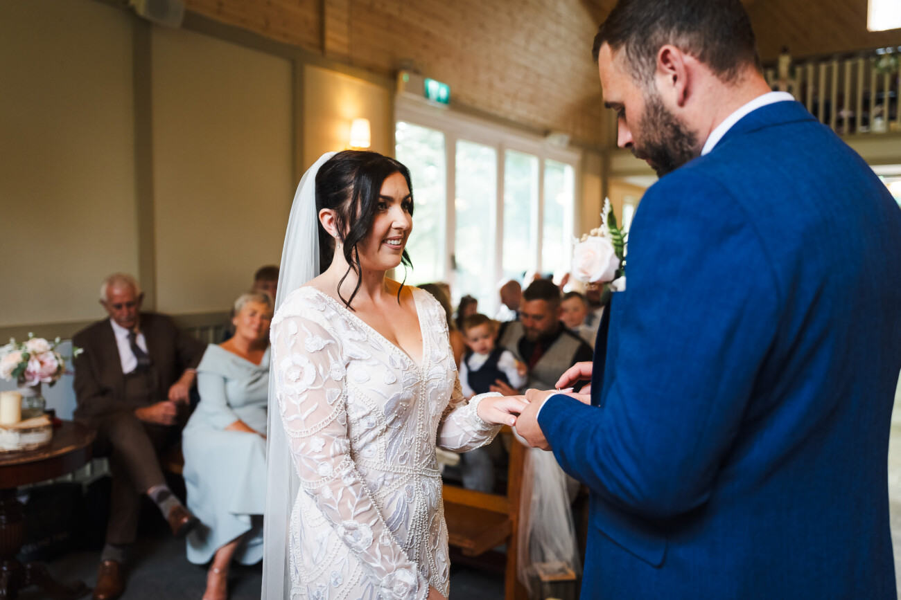 A Relaxed Summer Wedding at Village Barn, Tyrellspass | Amy & Eoghan 41 A bride and groom exchange rings during a wedding ceremony in a room with guests seated in the background.