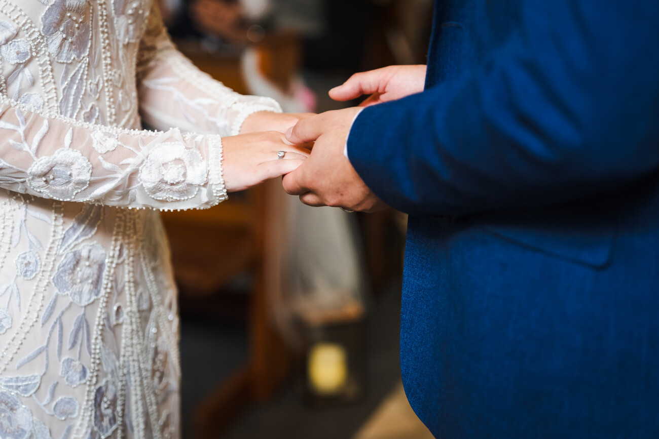 A Relaxed Summer Wedding at Village Barn, Tyrellspass | Amy & Eoghan 42 A person in a white lace dress and a person in a blue suit hold hands, with focus on their hands during a ceremony.